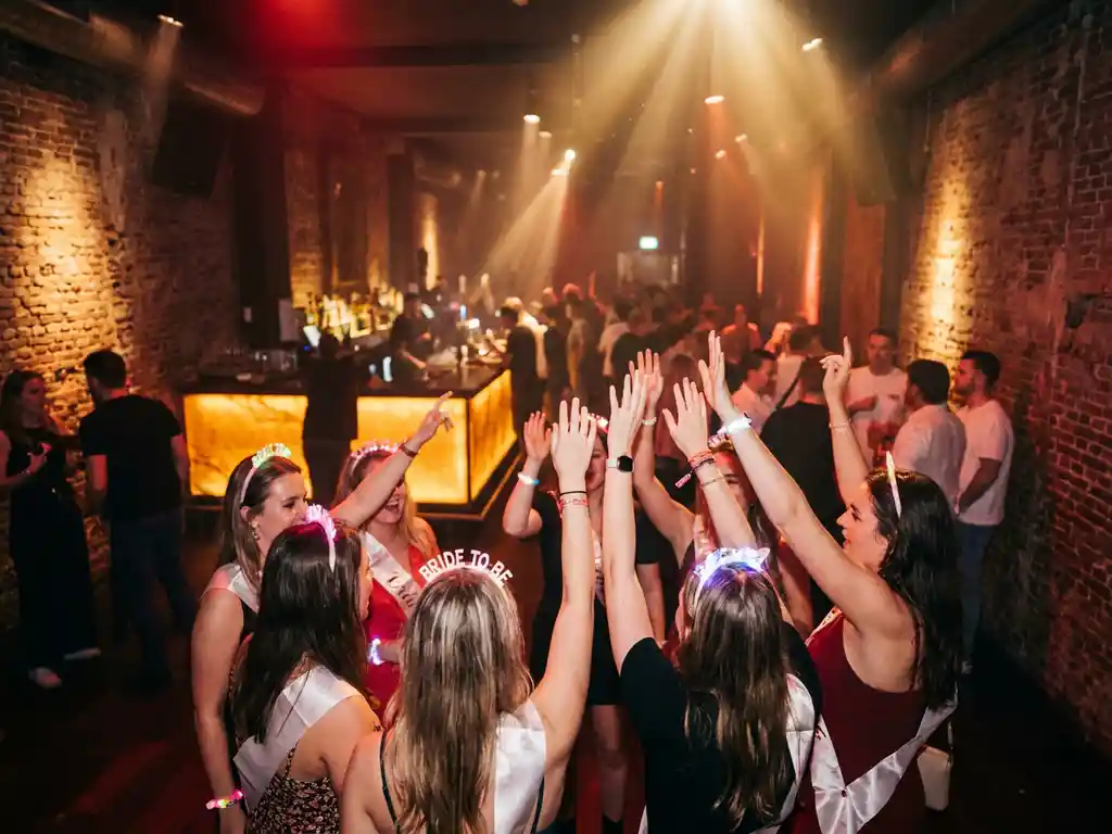 Vrijgezellenfeest groep danst in Amsterdams nachtclub interieur met bakstenen muren, amberkleurige bar en warme podiumverlichting.