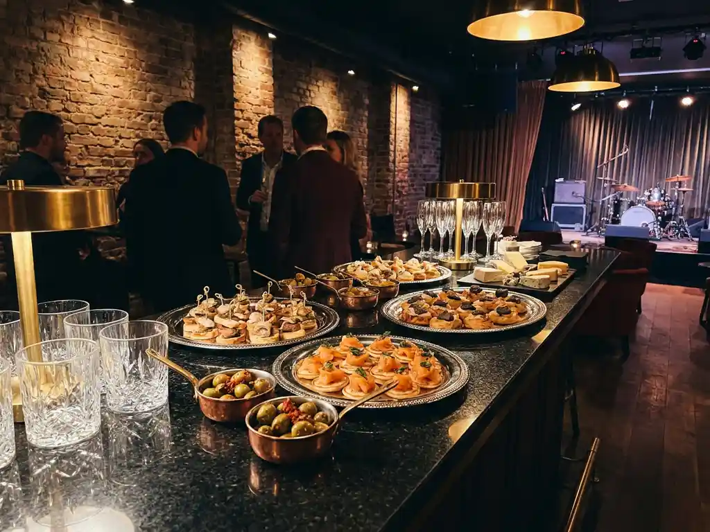 Luxe cateringbuffet op zwarte bartafel in Amsterdamse muziekclub, met zilveren schalen, hapjes en elegant glaswerk onder warm amberkleurig licht.