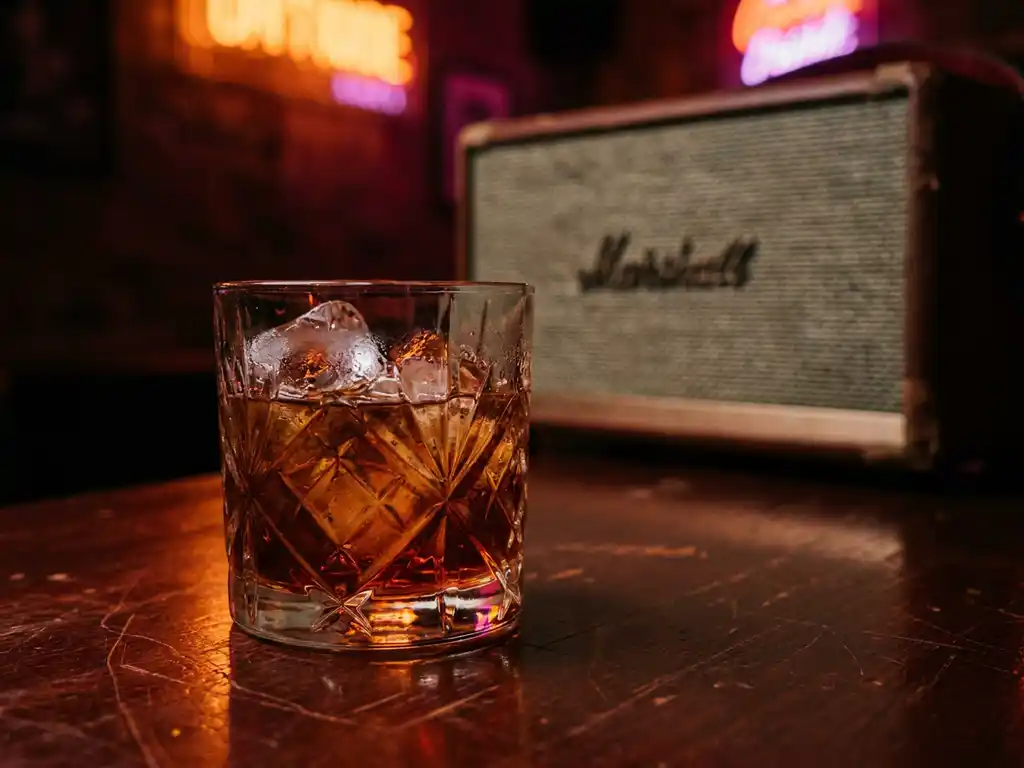 Amber cocktail met ijsblokjes op donker houten bar, oranje en magenta neongloed, condensatie op glas, intieme clubsfeer.