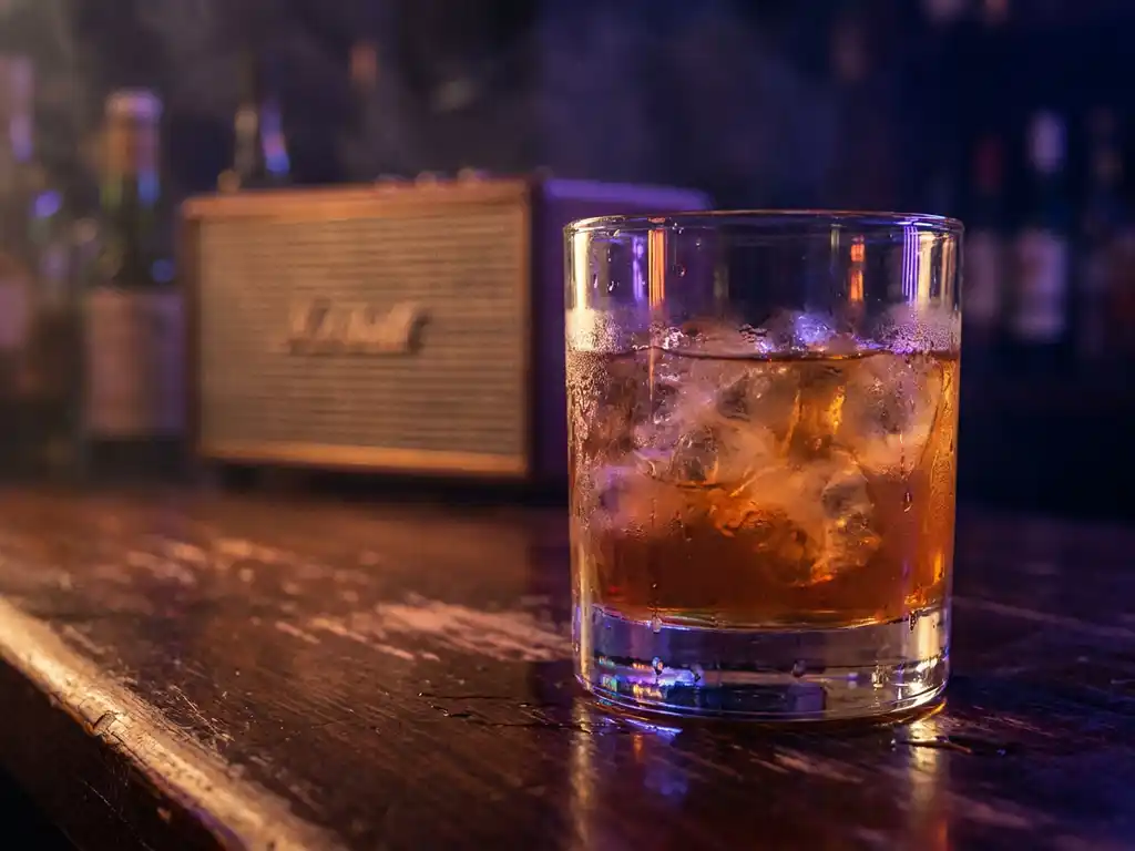 Beslagen cocktailglas met ijs op donker houten bar, verlicht door amber en violet neonlicht in intieme clubsfeer.