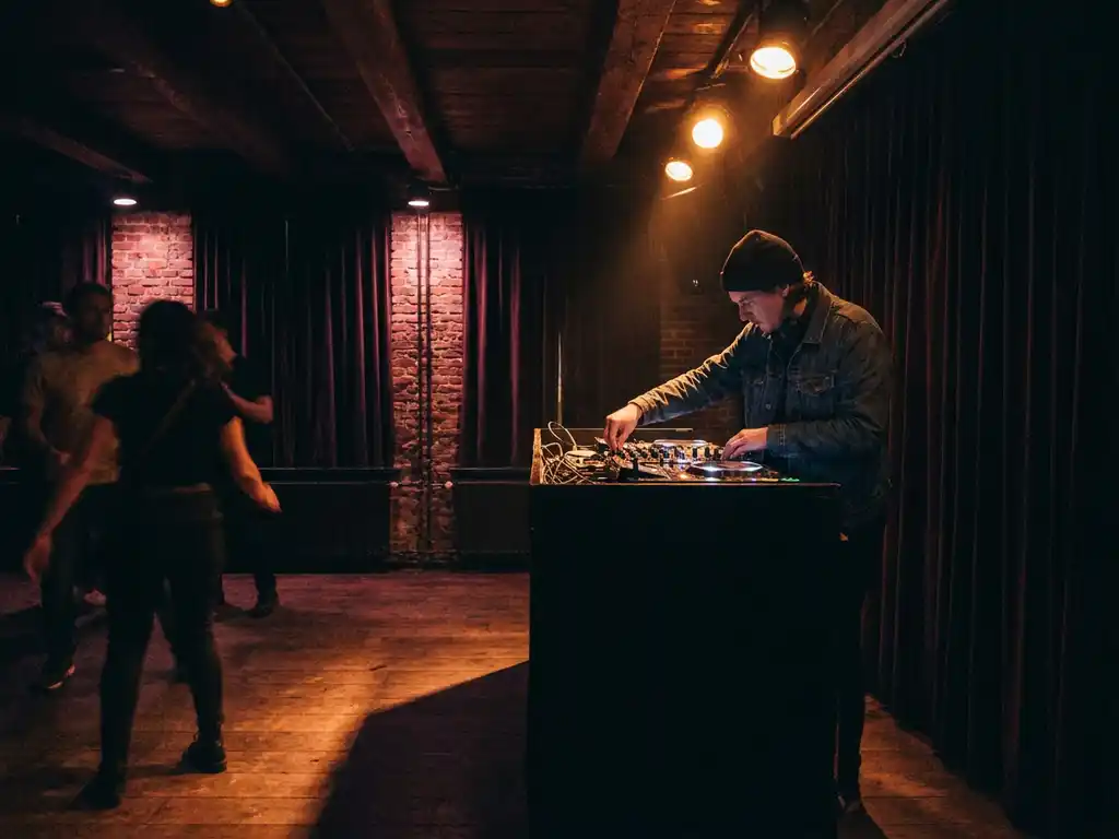 DJ achter de mixer in een intiem Amsterdams club met bakstenen muren, warm amberkleurig podiumlicht en houten dansvloer.