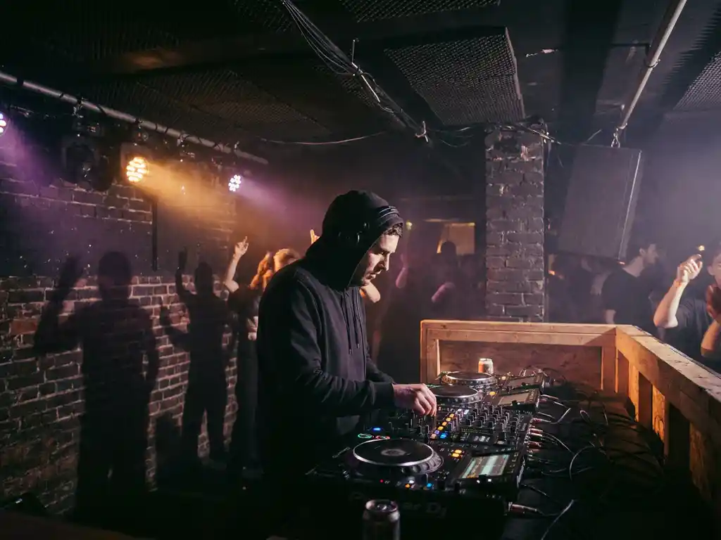 DJ achter professionele mixer op intiem podium, omgeven door amber en paars licht boven dansende menigte in underground club.