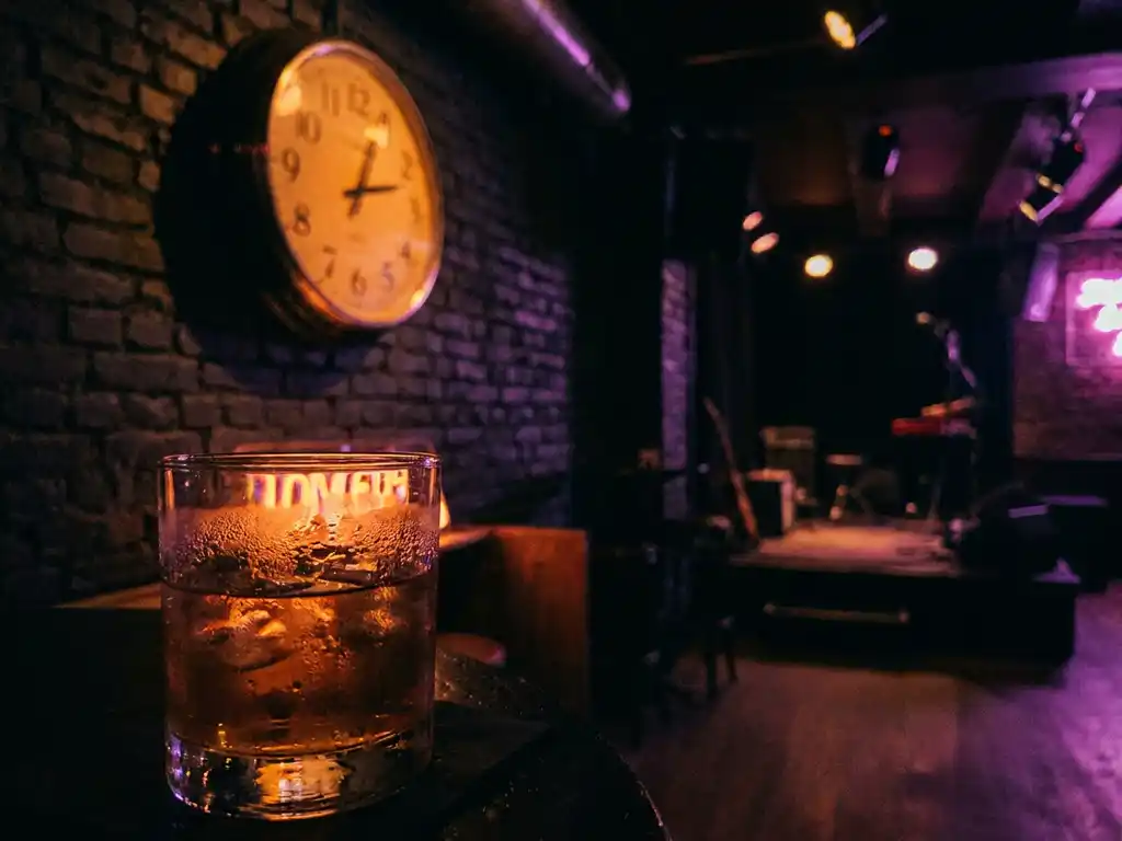 Vintage analoge klok op bakstenen muur van Amsterdams muziekcafé, met cocktailglas in amberkleurig neonlicht op de voorgrond.