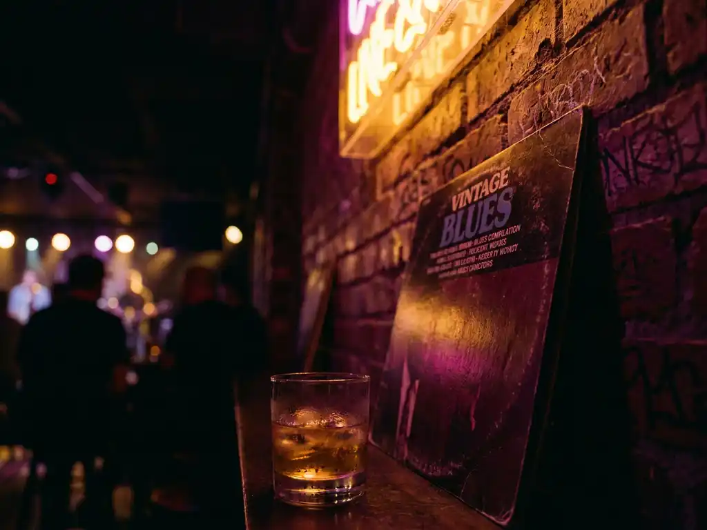 Vintage vinylplaat tegen verweerde bakstenen muur in Amsterdams jazzclub, met whiskey op ijs in amberkleurig neonlicht.