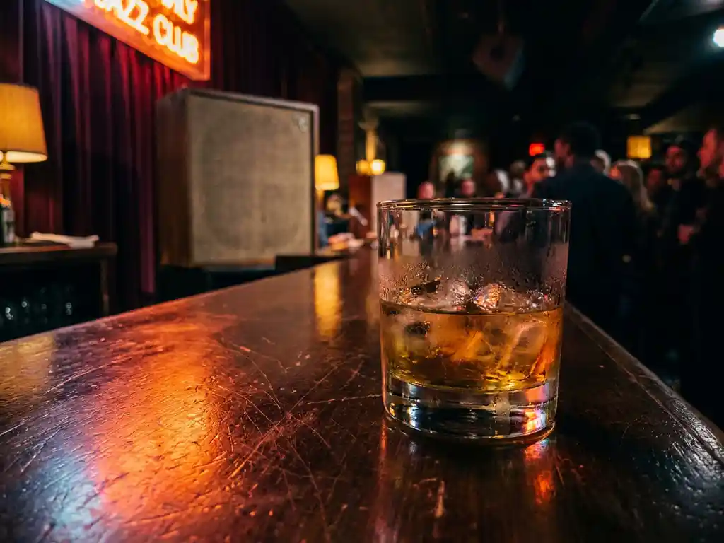 Whiskeyglas met ijs op donker houten bar, amberkleurig licht en neonreflecties, intieme clubsfeer met silhouetten op de achtergrond.