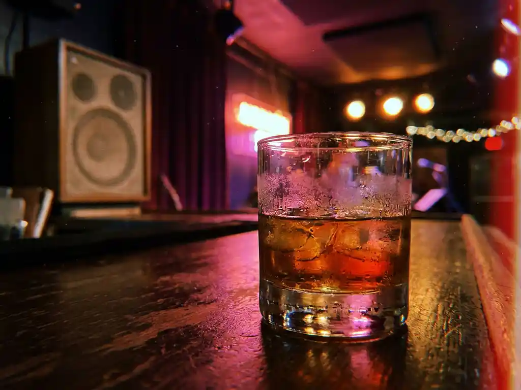 Condenserende whiskyglas met smeltend ijs op donker houten bar, verlicht door magenta en oranje neonlicht in intieme clubsfeer.