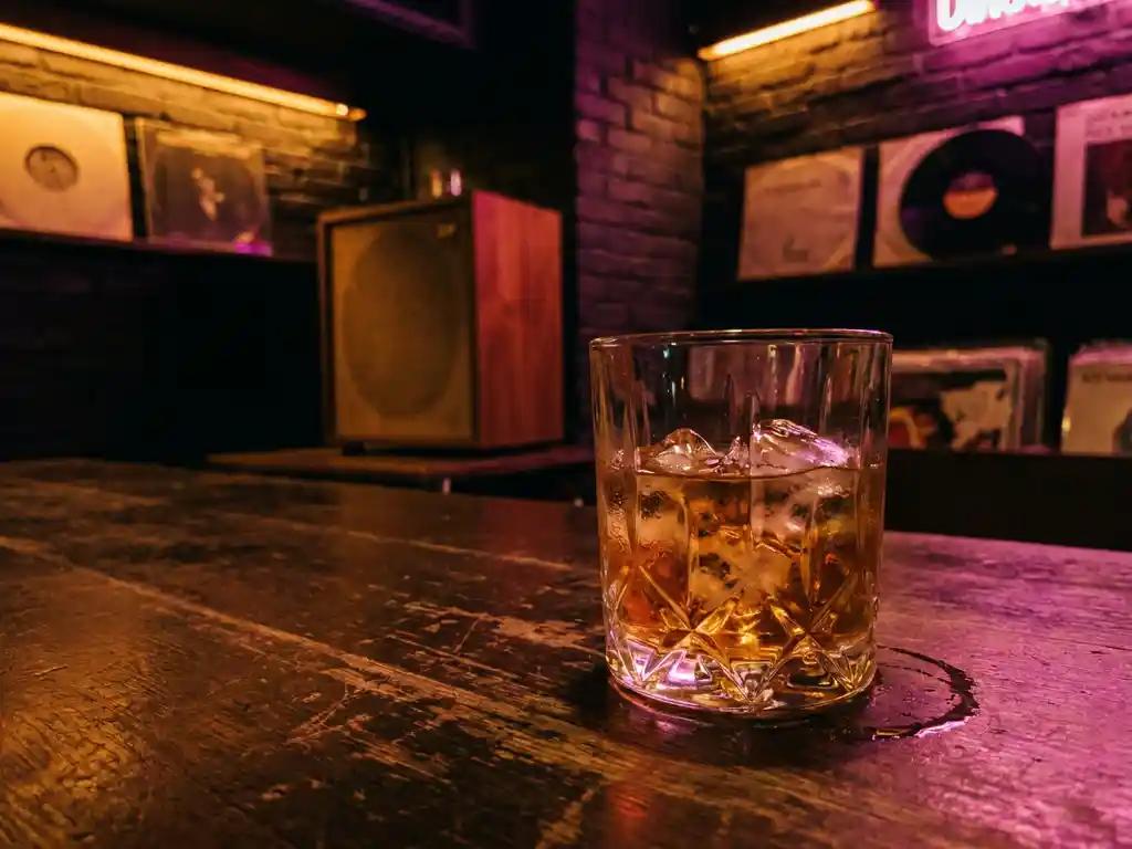 Vintage whiskyglas met ijsblokjes op versleten houten bar, omgeven door warm amber en magenta neonlicht in intieme underground jazzclub.